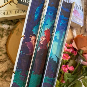 May include: Close-up of three hardcover books with illustrated spines. The spines feature fantasy art with figures in a forest setting, in shades of blue, purple, and green. The book titles are partially visible.