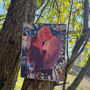 May include: A rectangular art print depicts a romantic scene of a couple embracing. The man has blonde hair and wears a light pink shirt, while the woman has red hair and a dark purple dress. The background features a blue and white abstract design.