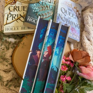 May include: A stack of fantasy novels with gold accents and a gold crown. The book spines feature illustrations of characters in a forest setting. The titles include "The Cruel Prince" and "The Queen of Nothing".