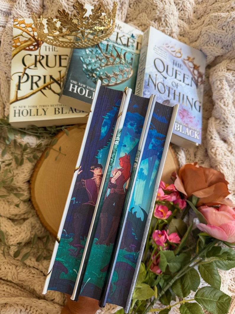 May include: A stack of fantasy novels with gold accents and a gold crown. The book spines feature illustrations of characters in a forest setting. The titles include "The Cruel Prince" and "The Queen of Nothing".