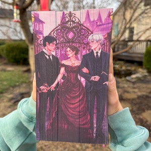 Op de afbeelding: Een houten boek met een fantasie-illustratie. Het kunstwerk toont drie figuren in formele kleding voor een smeedijzeren poort. Het kleurenpalet omvat tinten paars, zwart en rood.