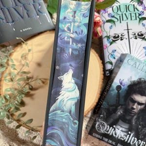 May include: A book with a dark cover featuring the title "Quicksilver" and a wolf illustration. The spine displays a detailed design with a wolf, a sword, and a butterfly. Other books are visible in the background.