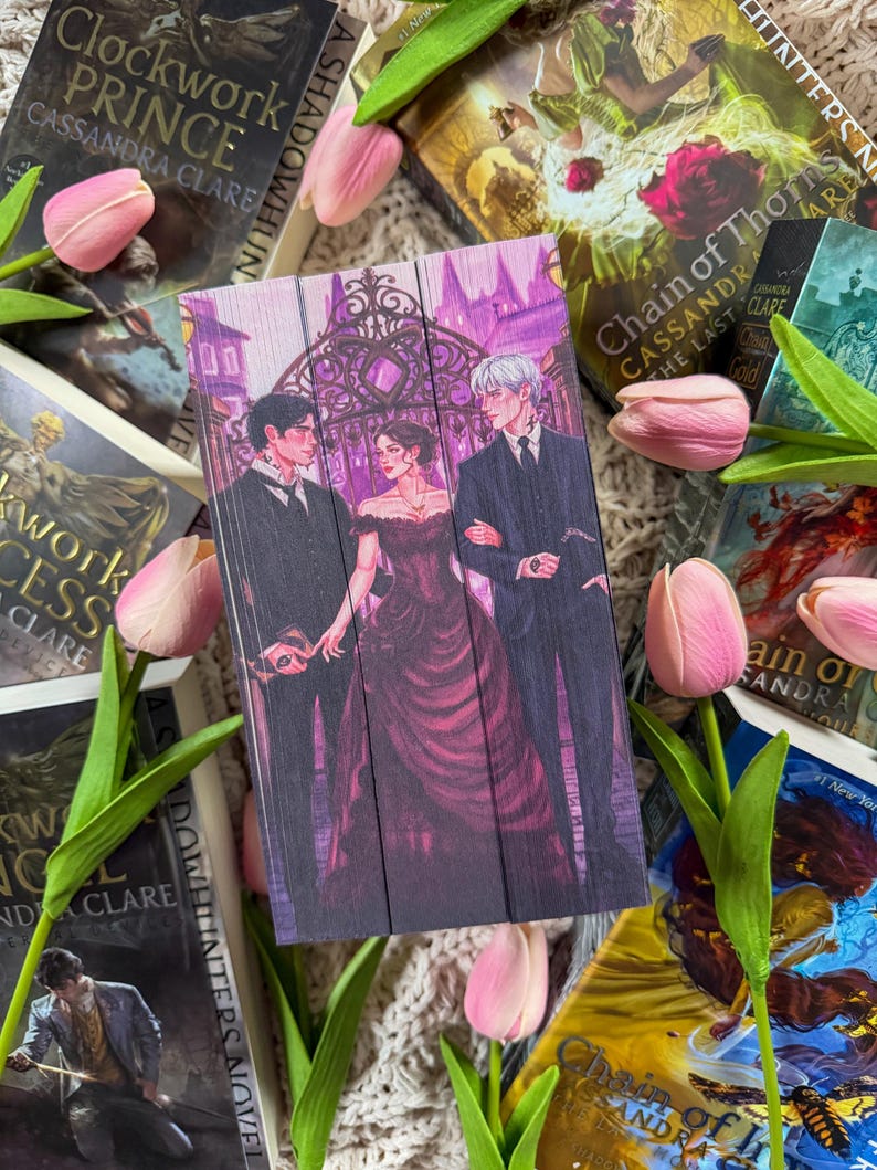 Op de afbeelding: Een houten boekenlegger met een romantische illustratie van drie figuren in formele kleding, tegen een achtergrond van een smeedijzeren poort. De boekenlegger is omgeven door boeken en roze tulpen, wat een literair thema cre&euml;ert.