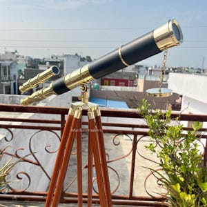 May include: A brass and black telescope mounted on a wooden tripod. The telescope has a long barrel with gold accents and a small secondary scope. The tripod has three brown wooden legs. The text "Maritimemakers" is visible on the tripod.