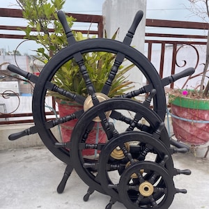 May include: A set of four black wooden ship wheels of varying sizes, with brass center hubs. The wheels are arranged in a row, with the largest in the back and the smallest in the front. The wheels are decorative nautical items.