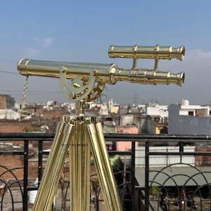 May include: A brass telescope on a tripod, with a cityscape and blue sky in the background. The telescope features a long barrel with a chain and a smaller, parallel scope. The tripod has three legs.