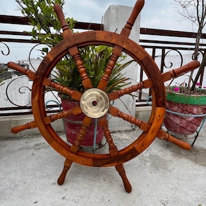 May include: A large, wooden ship's wheel with a brass center. The wheel is a rich, dark brown color and has multiple spokes and handles. It is standing on a concrete surface, with plants in the background.