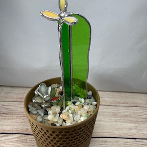May include: A stained glass cactus sculpture in a brown pot. The cactus is green with a yellow and white flower at the top. The pot is filled with small stones and a small glass succulent.