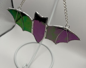 Puede incluir: Un murciélago de vidriera con alas verdes y moradas, delineado en plata. El murciélago está suspendido por una cadena plateada. Las alas tienen una cualidad reflectante e iridiscente. Es un artículo decorativo.