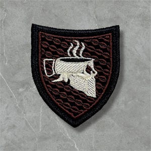 May include: Embroidered patch featuring a coffee cup with steam rising, set against a brown background with a textured pattern. The patch has a black border and a shield shape, suitable for jackets or bags.