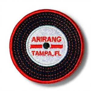 May include: Embroidered patch with a circular design. The patch features a white center with the word "ARIRANG" above "TAMPA, FL" in red text. The outer ring is red, and the middle ring is navy blue.