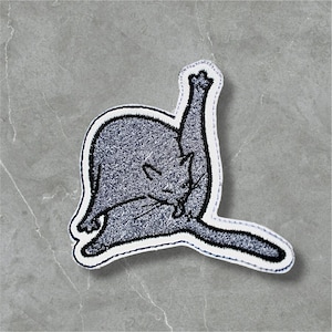 Op de afbeelding: Een grijze kattenpatch met een witte rand, geborduurd met zwart garen. De kat wordt afgebeeld terwijl hij zich verzorgt, met één poot omhoog. De patch staat op een grijze achtergrond.