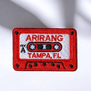 May include: A red embroidered patch in the shape of a cassette tape. The patch features the text "ARIRANG TAMPA, FL" in white, with a black and white design in the center. The patch has a white background.