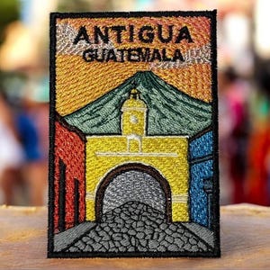 May include: Embroidered patch featuring the words "ANTIGUA GUATEMALA" above a colorful depiction of a building with an archway, a mountain, and colorful walls. The patch is rectangular with a black border and is likely a souvenir or travel-themed item.