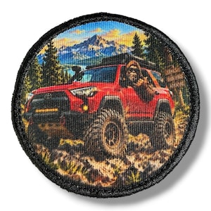 May include: A circular embroidered patch featuring a red off-road vehicle driving through a mountainous forest landscape. A brown, furry creature is visible in the driver seat. The patch has a thick black stitched border and a detailed graphic.