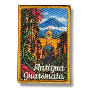 May include: A rectangular embroidered patch with a gold border featuring a scenic view of Antigua Guatemala. The design includes a yellow archway, a blue volcano, green foliage, pink flowers, and a quetzal bird. The text reads Antigua Guatemala.