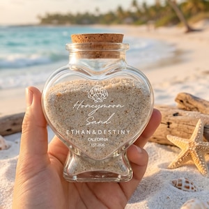 Puede incluir: Una botella de vidrio en forma de corazón llena de arena, etiquetada como "Honeymoon Sand" con nombres y una fecha. La botella tiene un tapón de corcho. El fondo muestra una playa con conchas marinas y una estrella de mar.