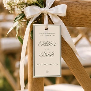 Può includere: Un cartoncino rettangolare color crema con bordo nero, legato a una sedia di legno con un nastro di raso bianco e un bouquet di fiori bianchi. Il cartoncino riporta la scritta: THIS SEAT IS KINDLY RESERVED FOR THE Mother of the Bride MARGARET WINSLOW.