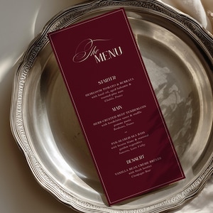 Może przedstawiać: Ciemnobordowa, prostokątna karta menu z białym tekstem szeryfowym i skryptowym zatytułowana The Menu, zawierająca sekcje Starter, Main i Dessert, umieszczona na okrągłym, srebrzystym metalowym talerzu z ozdobnym, falistym brzegiem.