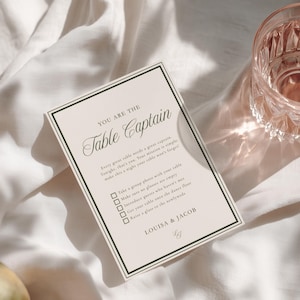 May include: A rectangular wedding table card with a cream background and black double-line border. The text reads: YOU ARE THE Table Captain, followed by instructions and a checklist. The card is signed LOUISA & JACOB at the bottom with a small logo.