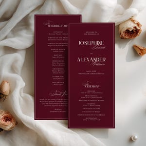 May include: Two burgundy wedding programs with gold trim, featuring elegant calligraphy. The programs are set against a backdrop of soft white fabric, dried roses, and pearls. The text includes names, ceremony details, and a thank you.