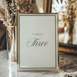 Puede incluir: Un letrero de número de mesa enmarcado en oro con fondo blanco. El texto "TABLE Three" está escrito en negro. El letrero está sobre una superficie de mármol. Flores secas y un jarrón son visibles en el fondo.