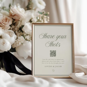 May include: A framed sign with the text "Share your Shots" and a QR code, displayed on a satin surface. The sign is in a gold frame, and a bouquet of white and blush flowers is in the background.
