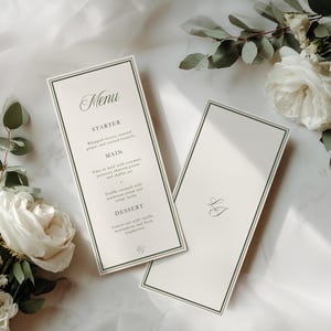 May include: Two ivory-colored menu cards with a dark green border and elegant script. One card displays a menu with headings for Starter, Main, and Dessert. The other card has initials. White roses and greenery are arranged around the cards.