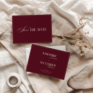 Può includere: Biglietti d'invito di nozze color bordeaux con testo bianco. Un biglietto recita "Save the Date" in elegante scrittura. L'altro biglietto mostra nomi e dettagli dell'evento. I biglietti sono su un panno beige con una piccola candela e grano secco.