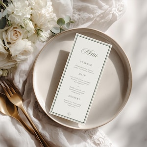 May include: A menu card with a dark green border sits on a cream-colored plate, next to gold-colored silverware. The menu lists starter, main, and dessert courses. White flowers and greenery are in the background.