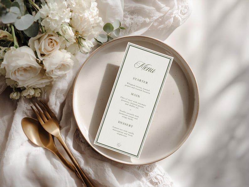 May include: A menu card with a dark green border sits on a cream-colored plate, next to gold-colored silverware. The menu lists starter, main, and dessert courses. White flowers and greenery are in the background.