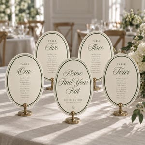 Può includere: Cinque segnaposto ovali color bianco sporco con bordi verde scuro, ciascuno montato su un piccolo supporto in metallo color ottone. Le carte mostrano testo nero che include numeri di tavolo, nomi degli ospiti e la scritta Please Find Your Seat.