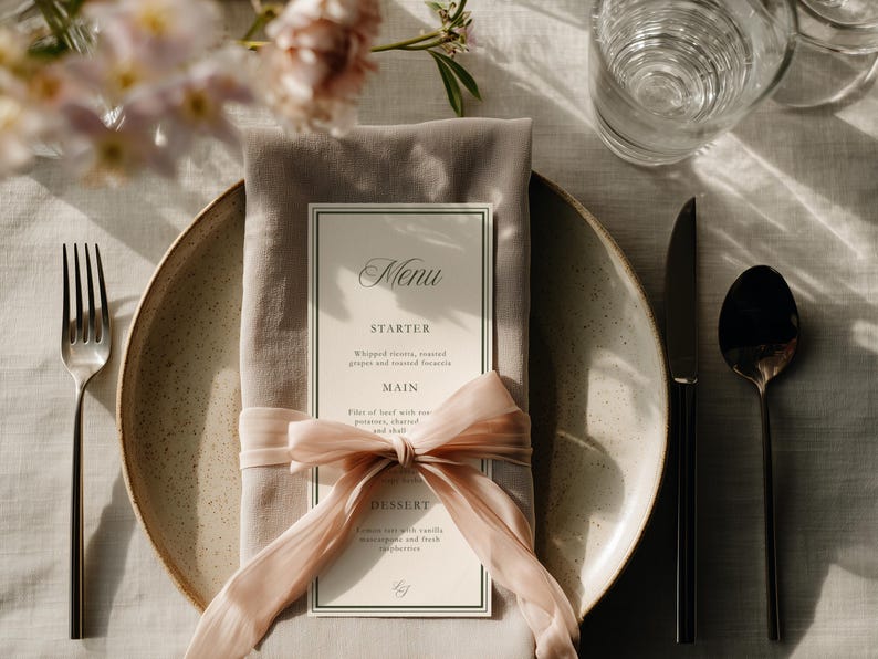 May include: A formal table setting with a menu card, napkin, and silverware. The menu card is cream-colored with black text and a decorative border. A pink ribbon is tied around the napkin. A fork, knife, and spoon are arranged on the table.