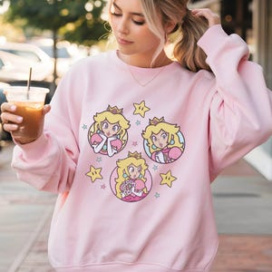 May include: A light pink sweatshirt featuring three cartoon images of Princess Peach, each in a circular frame. The princess has blonde hair, a crown, and a pink dress. Yellow stars are scattered around the images.