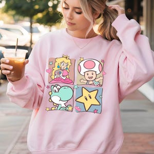 May include: A light pink sweatshirt featuring a four-panel design with cartoon characters. The panels include Princess Peach, Toad, Yoshi, and a yellow star. The sweatshirt has long sleeves and a crew neck.