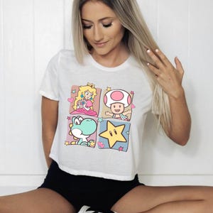 May include: White t-shirt featuring a colorful graphic of four characters from the Super Mario video game series. The design includes Princess Peach, Toad, Yoshi, and a yellow star, each in a separate square.