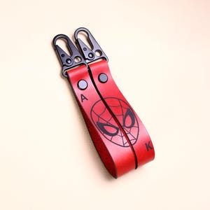 May include: Two red leather keychains with black metal clasps. Each keychain features a black Spider-Man mask design and the letters "A" and "K". The keychains are connected and lie on a light beige surface.