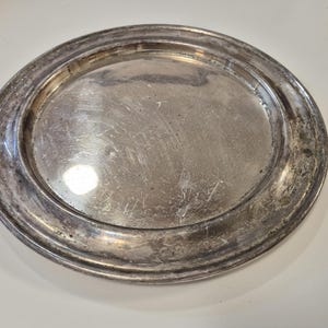 May include: A circular, silver-coloured metal tray with a broad, raised rim. The tray has a slightly oval form and shows signs of wear, with scratches and discolouration across the surface. The rim has a decorative edge.