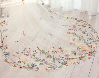May include: A sheer, ivory-colored wedding veil with delicate floral embroidery. The veil is adorned with colorful embroidered flowers in shades of pink, red, yellow, and green, creating a whimsical and romantic design. The veil is spread out on a light-colored wooden surface.