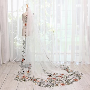 Pode incluir: Um longo véu de noiva branco com bordado floral. O véu apresenta um delicado design de flores e folhas laranja, vermelhas e verdes ao longo das bordas. O véu é exibido em um manequim em frente a uma janela.