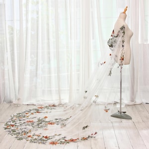 May include: A sheer white wedding veil with floral embroidery. The veil is displayed on a mannequin, showcasing the delicate design. The embroidery features orange, green, and white flowers and leaves, adding a touch of color and detail to the bridal accessory.