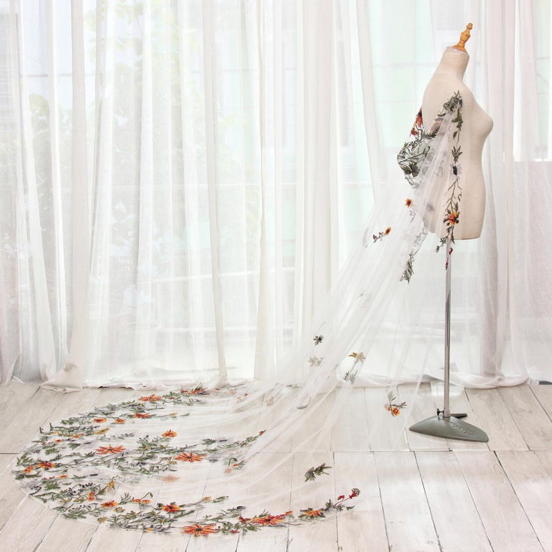 May include: A sheer white wedding veil with floral embroidery. The veil is displayed on a mannequin, showcasing the delicate design. The embroidery features orange, green, and white flowers and leaves, adding a touch of color and detail to the bridal accessory.