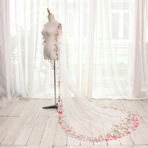 May include: A white bridal veil with floral embroidery. The veil is displayed on a mannequin, showcasing a delicate design of pink and green flowers along the edges and cascading down the side. The veil is long and flowing.