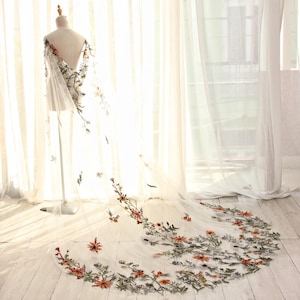 May include: A sheer white wedding veil with floral embroidery. The veil is displayed on a mannequin, with the floral design cascading down the length. The embroidery features orange and green flowers and leaves.