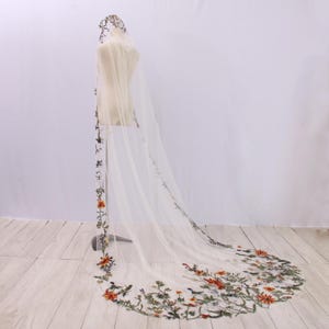 May include: A sheer, ivory-colored bridal veil with a floral embroidered design. The veil features a hood and a long train, with the embroidery in shades of orange, green, and white. The veil is displayed on a mannequin.