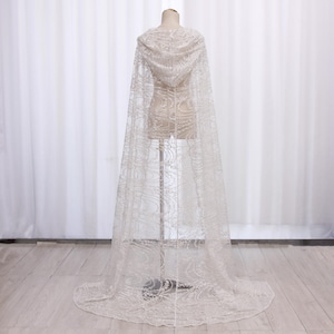 May include: An ivory lace hooded cape, featuring a detailed floral design. The cape is displayed on a mannequin, highlighting its flowing silhouette and delicate texture. The floor-length cape is suitable for special events and weddings.