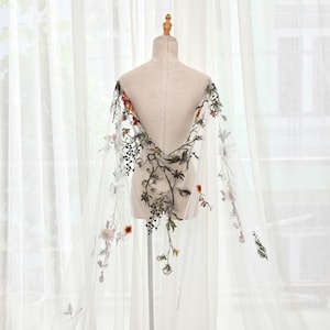 May include: A sheer white cape with intricate floral embroidery in shades of green, red, and yellow. The cape is draped over a mannequin, showcasing the delicate design and flowing fabric. The floral pattern is concentrated along the edges and center.