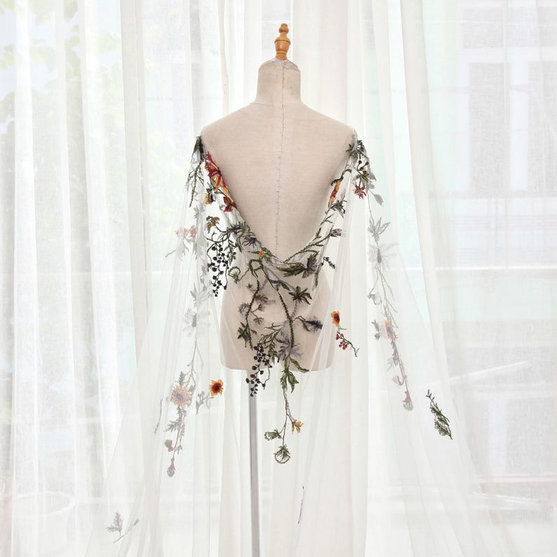 May include: A sheer white cape with intricate floral embroidery in shades of green, red, and yellow. The cape is draped over a mannequin, showcasing the delicate design and flowing fabric. The floral pattern is concentrated along the edges and center.