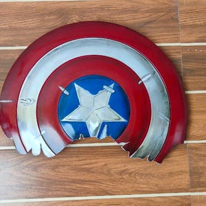 May include: A distressed, half-circle shield in red, white, and blue, featuring a central blue circle with a white star. The shield has a metallic silver trim and visible damage along the bottom edge, suggesting battle wear.
