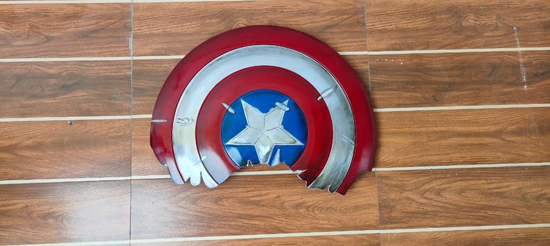 May include: A distressed, half-circle shield in red, white, and blue, featuring a central blue circle with a white star. The shield has a metallic silver trim and visible damage along the bottom edge, suggesting battle wear.